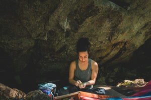 girl climber sitting on crash pads in front of boulder using skin salve after climbing on hands.