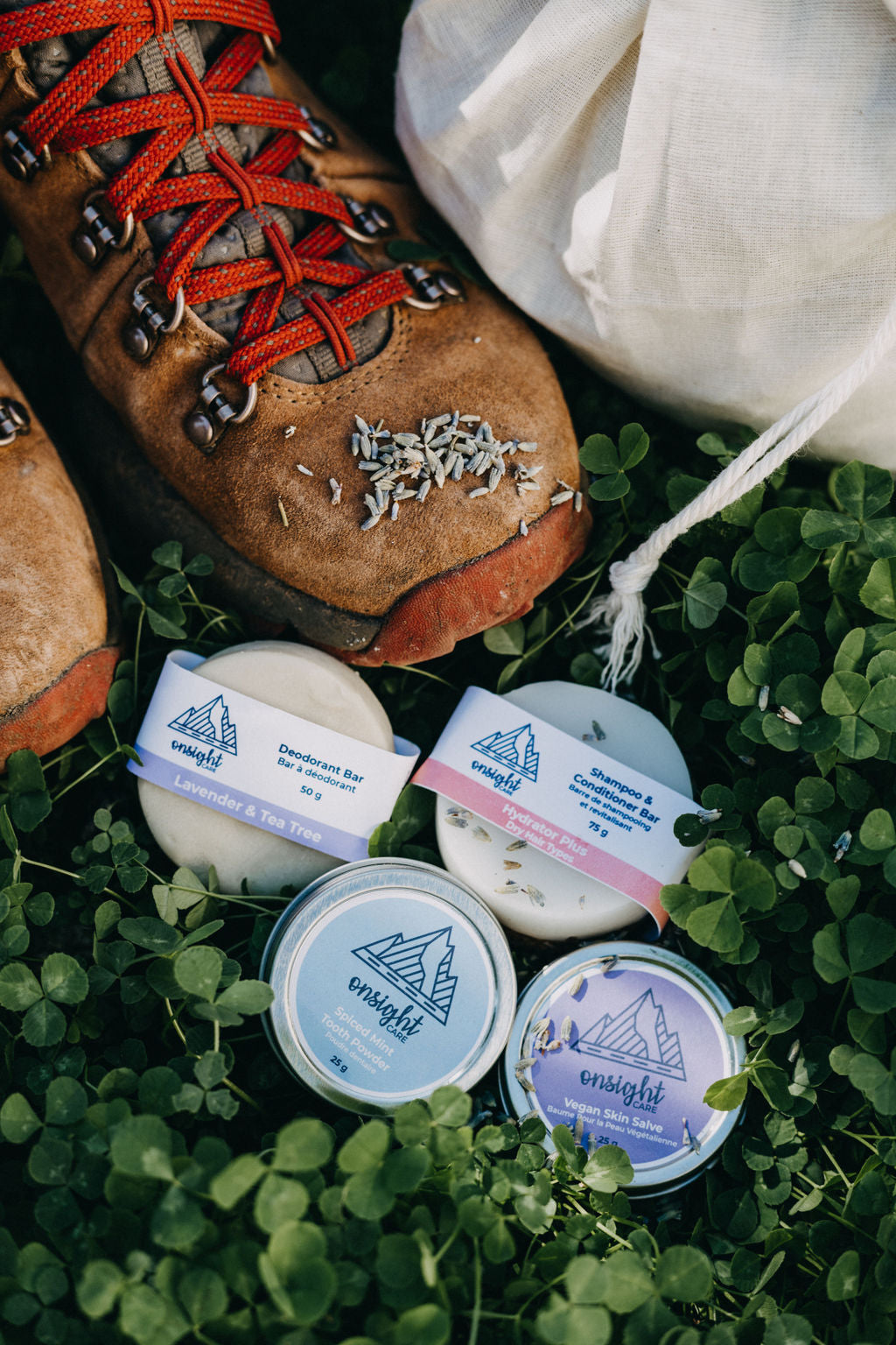 hiking boots with lavender sprinkled on top. Deodorant bar, shampoo & conditioner bar, tooth powder, vegan skin salve, and cotton bulk bag delicately sitting around hiking boots in the clover grass.